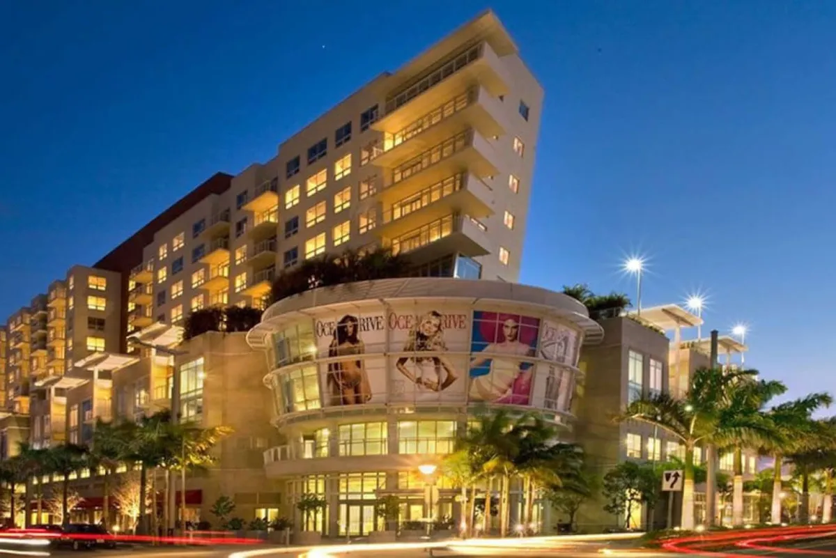 A contemporary exterior scene featuring stylish modern architecture, large promotional banners suggesting nearby shopping or cultural events, palm trees, and vibrant ambient lighting. The lively atmosphere hints at the urban energy and cultural richness of the area.