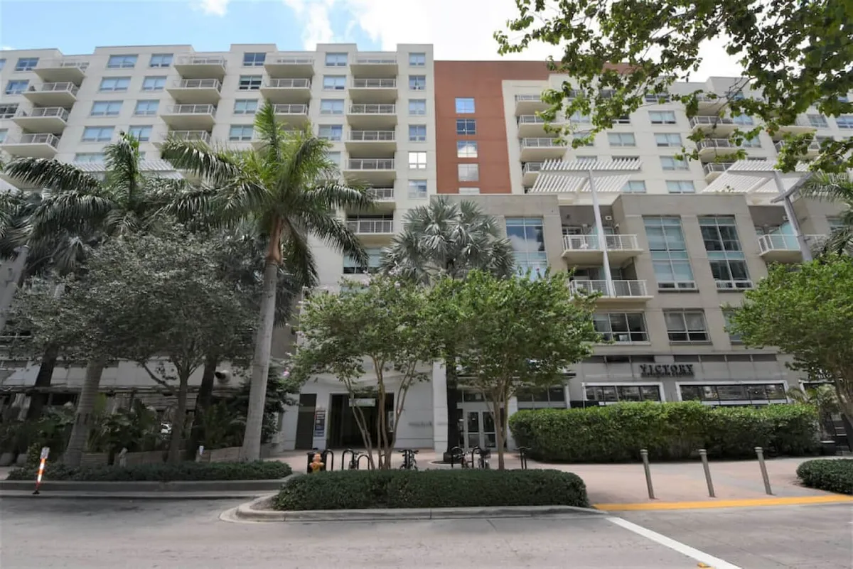 An exterior shot of a modern apartment building surrounded by lush greenery and featuring an inviting outdoor seating area. The scene conveys a welcoming, walkable neighborhood ambiance.
