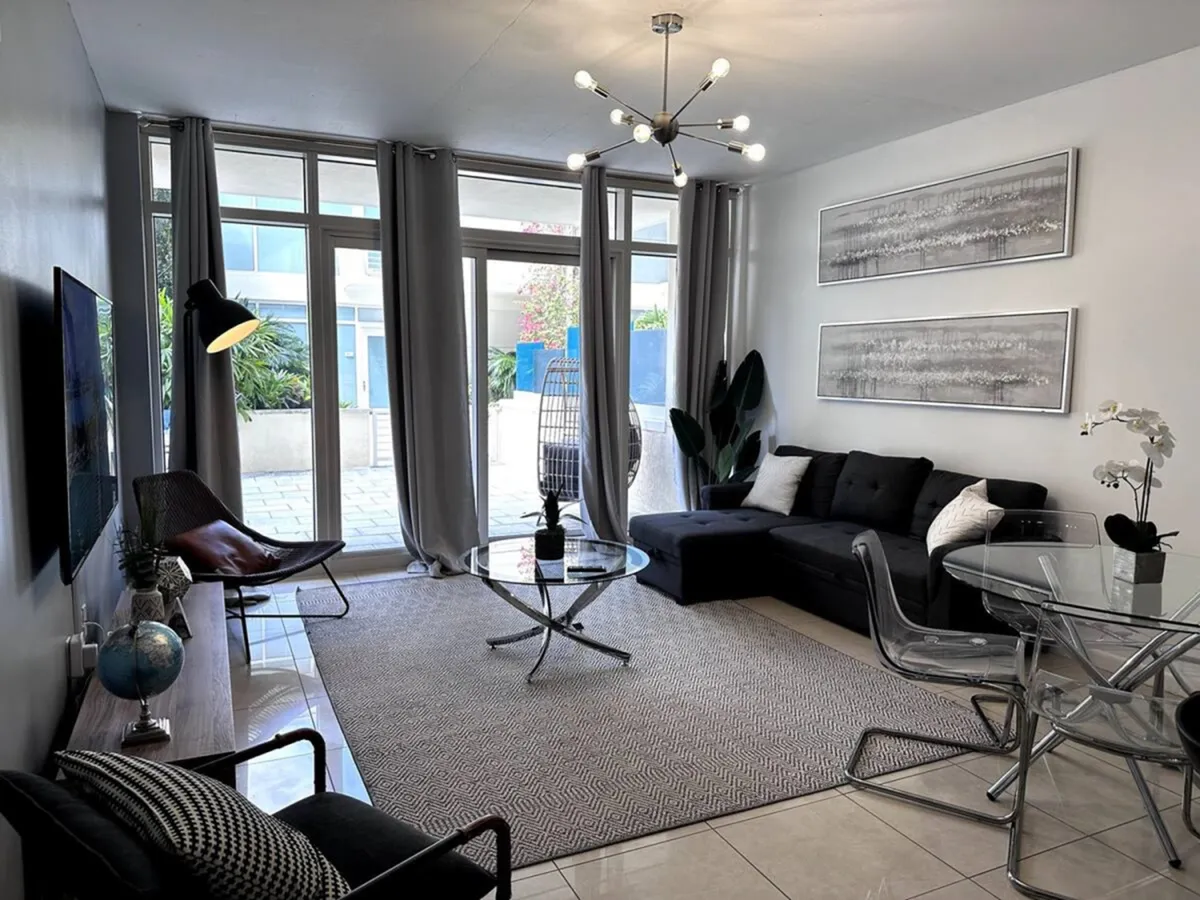 The image showcases a sunlit, modern living space with expansive floor-to-ceiling windows opening onto a private balcony. A plush black sectional sofa with colorful accent pillows invites relaxation, while a sleek glass coffee table and a striking chandelier add a touch of sophistication. The balcony offers sweeping city views, hinting at the vibrant Miami lifestyle just outside.