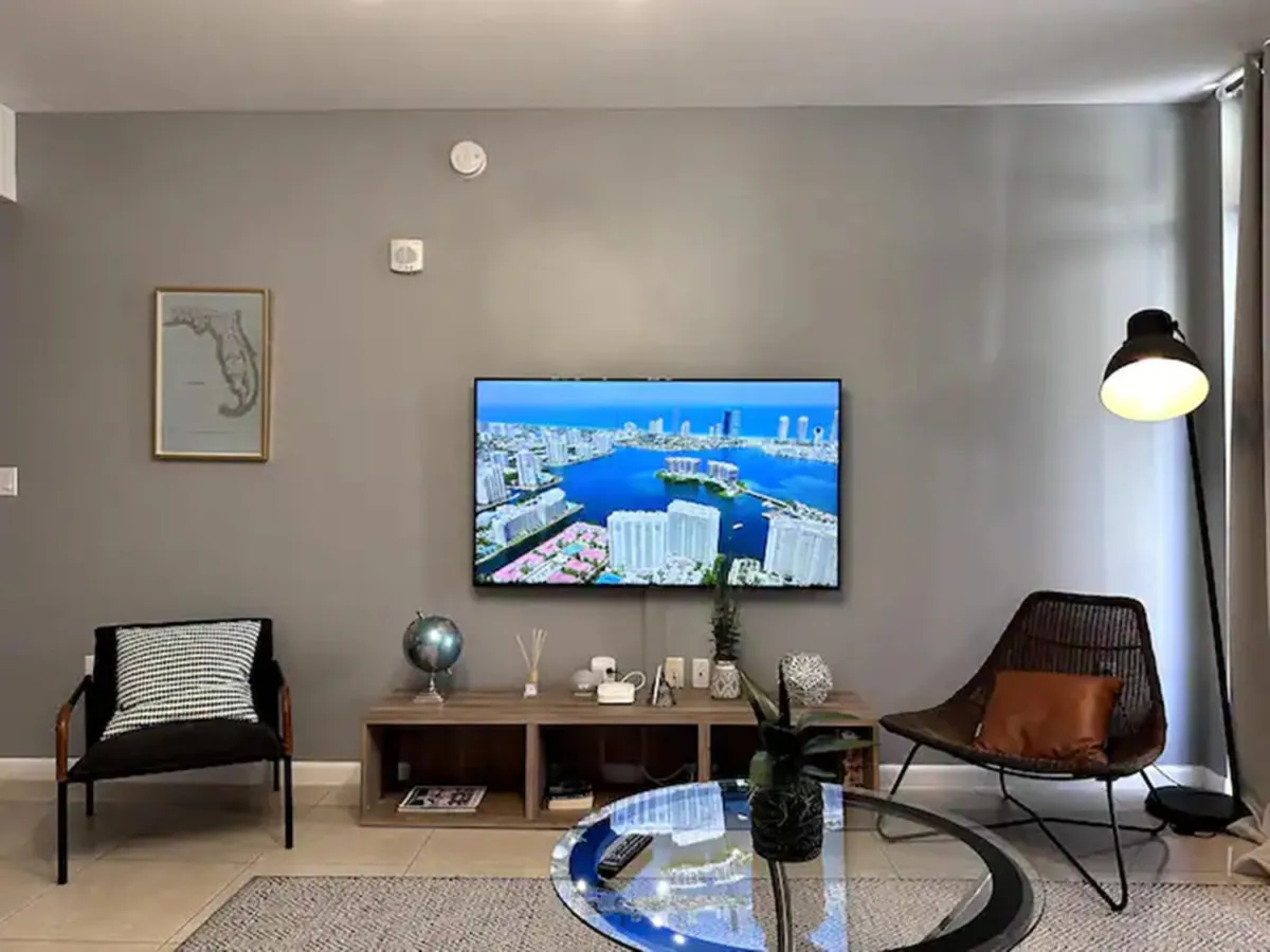This image features a modern living area with a wall-mounted flat screen TV displaying an aerial view of the city, a glass coffee table with a decorative plant, and two stylish chairs with cushions. A warm floor lamp and contemporary décor create a welcoming, social atmosphere ideal for relaxing or gathering before or after outings.