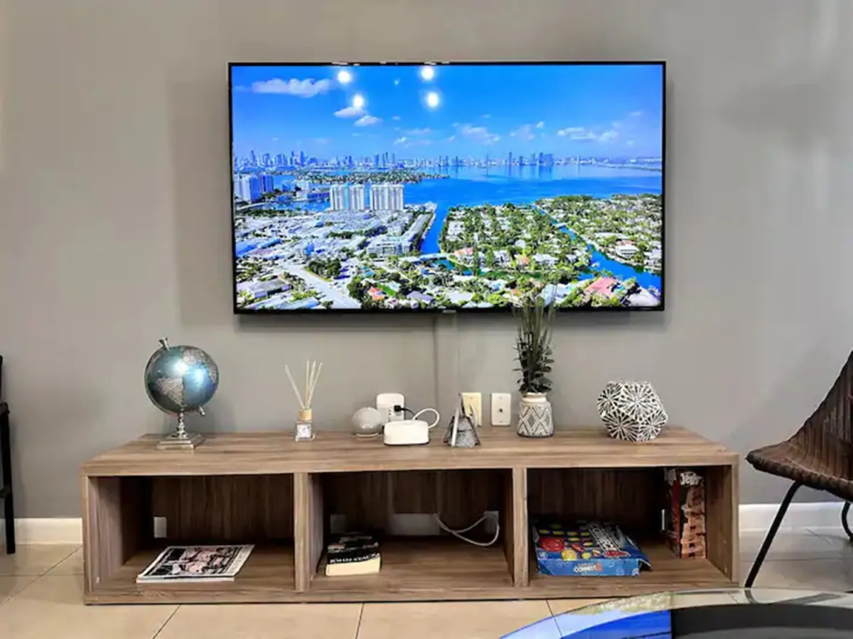 A calm, sophisticated living space is shown, with a large flat-screen TV featuring a vibrant cityscape and water view, a wooden media console with books, and a decorative globe. A modern chair peeks from the edge, hinting at group gatherings and trip planning.