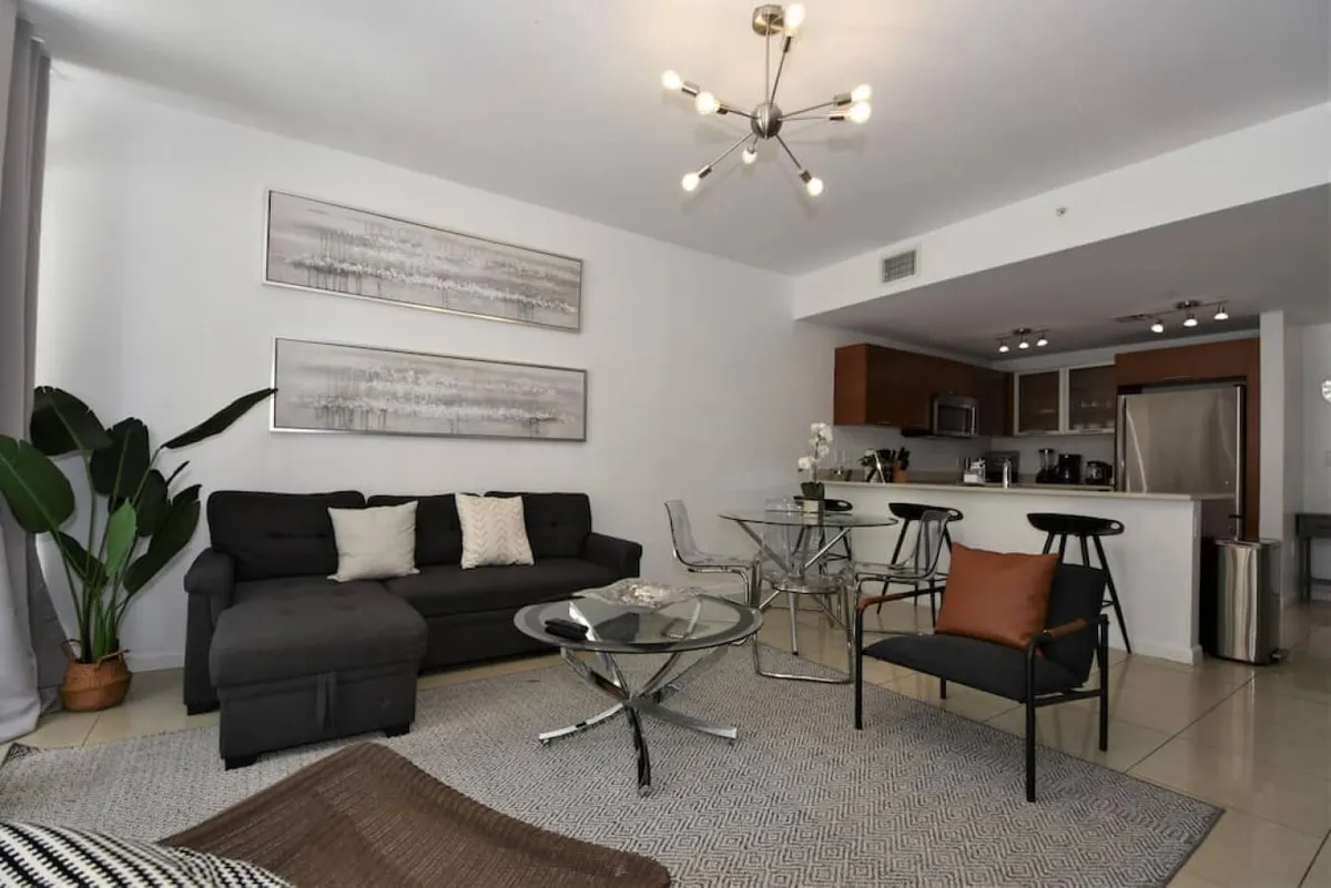 A spacious, stylish living and dining area with a dark sectional sofa adorned with cushions, a glass coffee table, and a modern chandelier. The open kitchen with a breakfast bar and stools is visible in the background, and large windows flood the space with natural light.