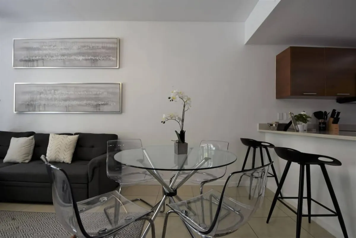 A contemporary living/dining zone featuring a sleek glass dining table with transparent chairs, a dark sofa with light throw pillows, and a kitchen counter with bar stools. Minimalist wall art adorns the space, and the overall design feels airy and inviting.