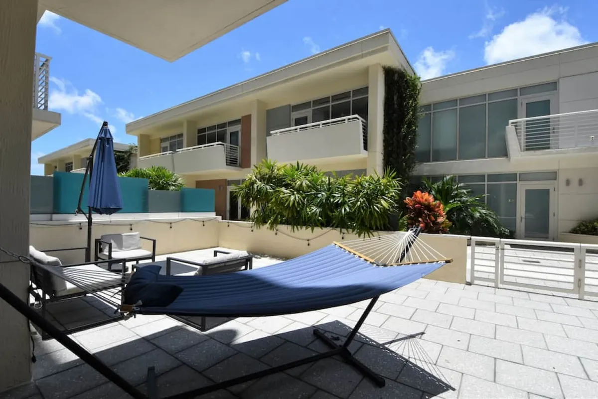 A serene outdoor patio features a blue hammock, inviting seating area with chairs and a table, a large umbrella for shade, and lush green landscaping, creating a peaceful corner within the city.