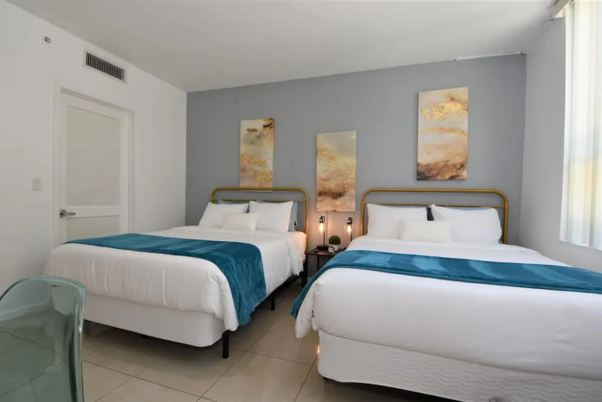 A bright, restful bedroom featuring two queen beds dressed in crisp white linens with teal throws, complemented by abstract gold and beige wall art above. A small bedside table hosts two modern lamps and a potted plant, while a translucent chair sits in the corner, and natural light streams in from a nearby window.