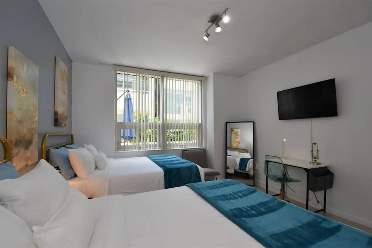 This image features a spacious bedroom with two queen beds dressed in teal blankets, a large window with vertical blinds letting in natural light, a wall-mounted TV, and a small desk with a transparent chair and mirror. The setting is calm and comfortable, inviting families to relax after a day exploring the city.