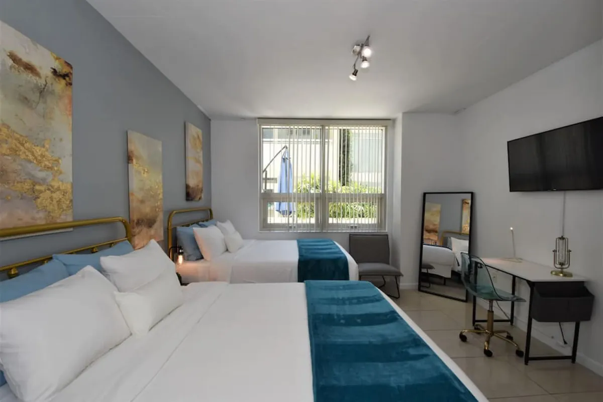 This image shows a similar angle as image-09, with two queen beds adorned in blue throws, a large window with blinds, a wall-mounted flat screen TV, and a desk area with a transparent chair and standing mirror. The room is bright and airy, ideal for resting between outings or preparing for the next family adventure.