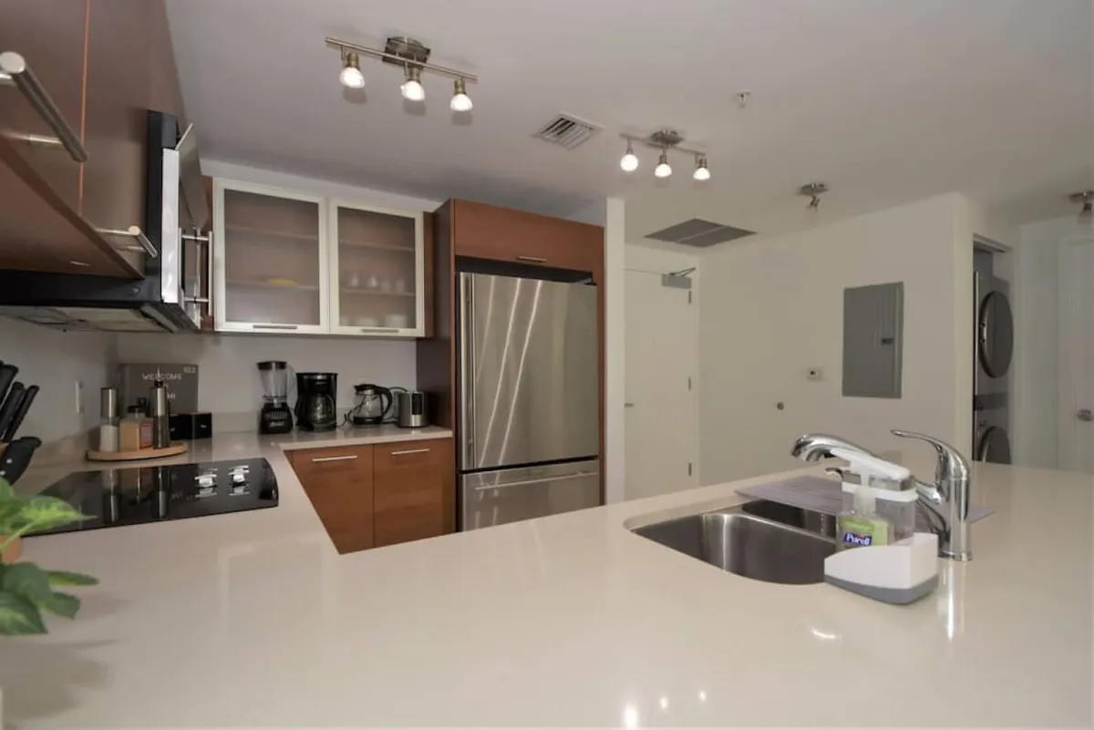 This angle shows the kitchen island with sink and soap dispenser, stainless steel refrigerator, coffee maker, and blender on the counter. In the background, a stacked washer and dryer hint at the unit's overall convenience. The space is bright, tidy, and designed for comfort and efficiency.