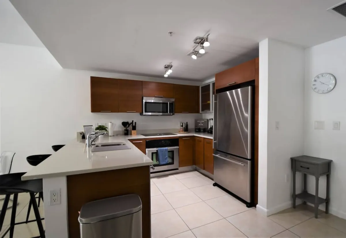 This image features a modern kitchen with stainless steel refrigerator, a microwave above the stove, a kitchen island with built-in sink and seating, and warm wooden cabinetry. The scene shows a layout conducive to meal preparation, casual dining, and conversation, with comfortable seating at the breakfast bar.