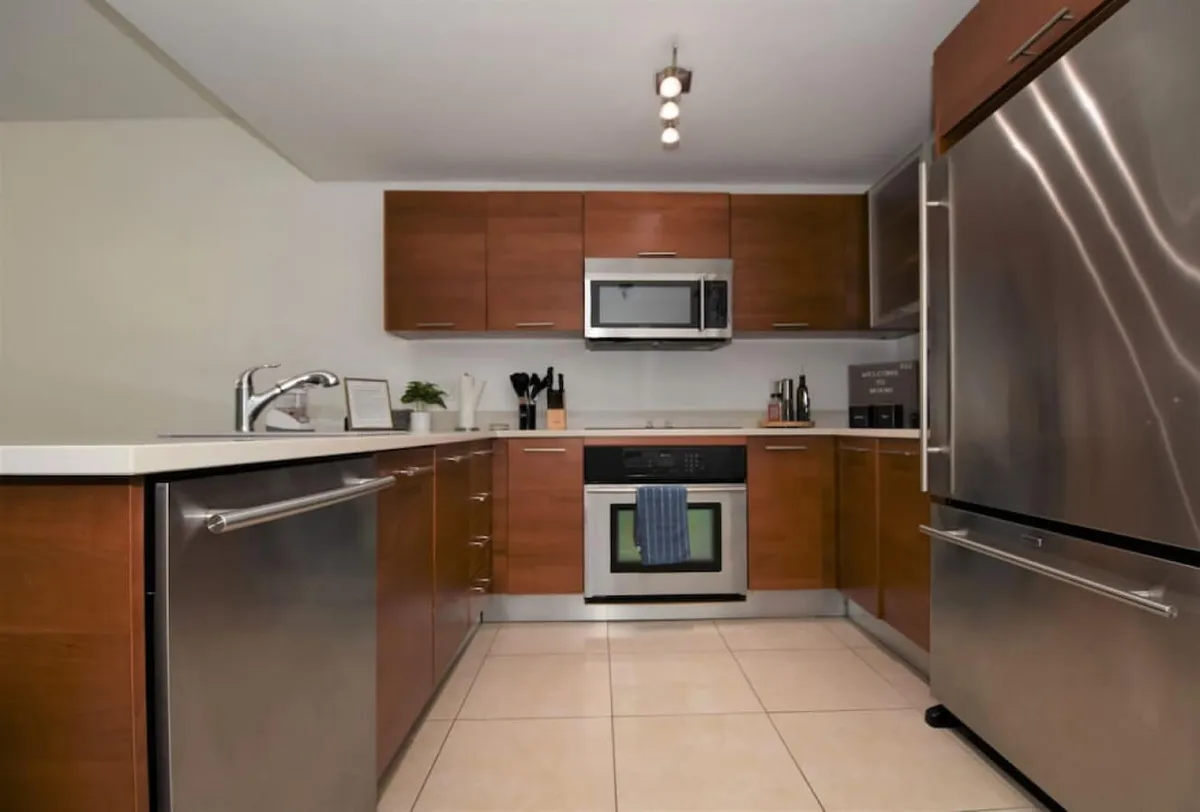 A closer look at the kitchen’s stainless steel refrigerator and oven, with a blue towel adding a pop of color. The microwave sits neatly above the oven, and the wooden cabinetry is sleek and modern. The workspace is organized, offering a practical and welcoming vibe for cooking meals or preparing drinks.