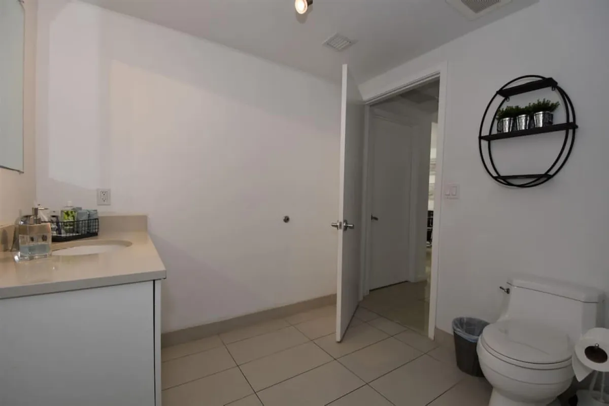 A view of a modern bathroom featuring a sleek toilet, a countertop with toiletries, and a circular wall shelf adorned with small plants. The open door reveals a glimpse into an adjacent room, adding a sense of flow and connection within the condo. The atmosphere is calm, functional, and subtly enhanced by decorative touches.