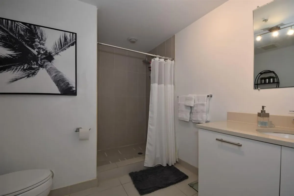 A modern bathroom with a clean shower area (with curtain), sleek vanity with sink and mirror, toilet, and striking black and white palm tree artwork. The tidy setup is both functional and visually appealing, with fresh towels and toiletries visible.
