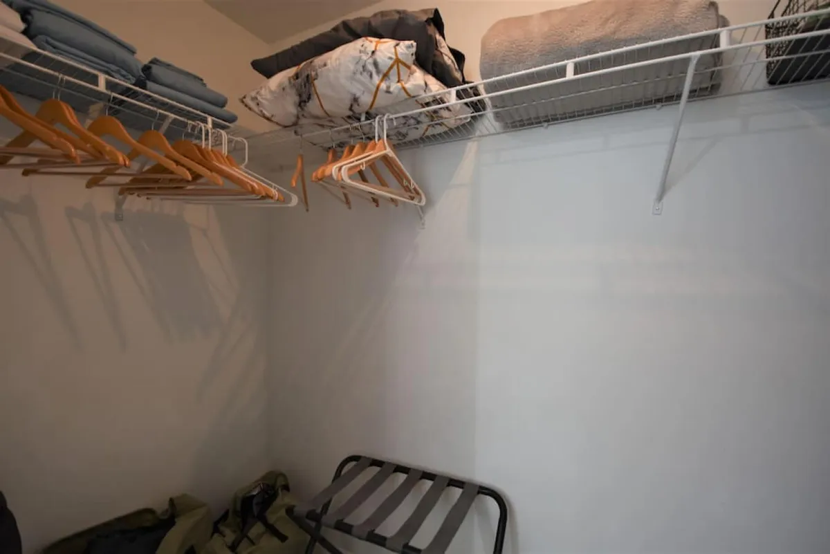 A well-organized closet space with wire racks holding hangers, neatly folded linens and pillows on the upper shelves, and a luggage rack at the bottom. The scene is practical and tidy, emphasizing storage solutions for guests and residents alike.