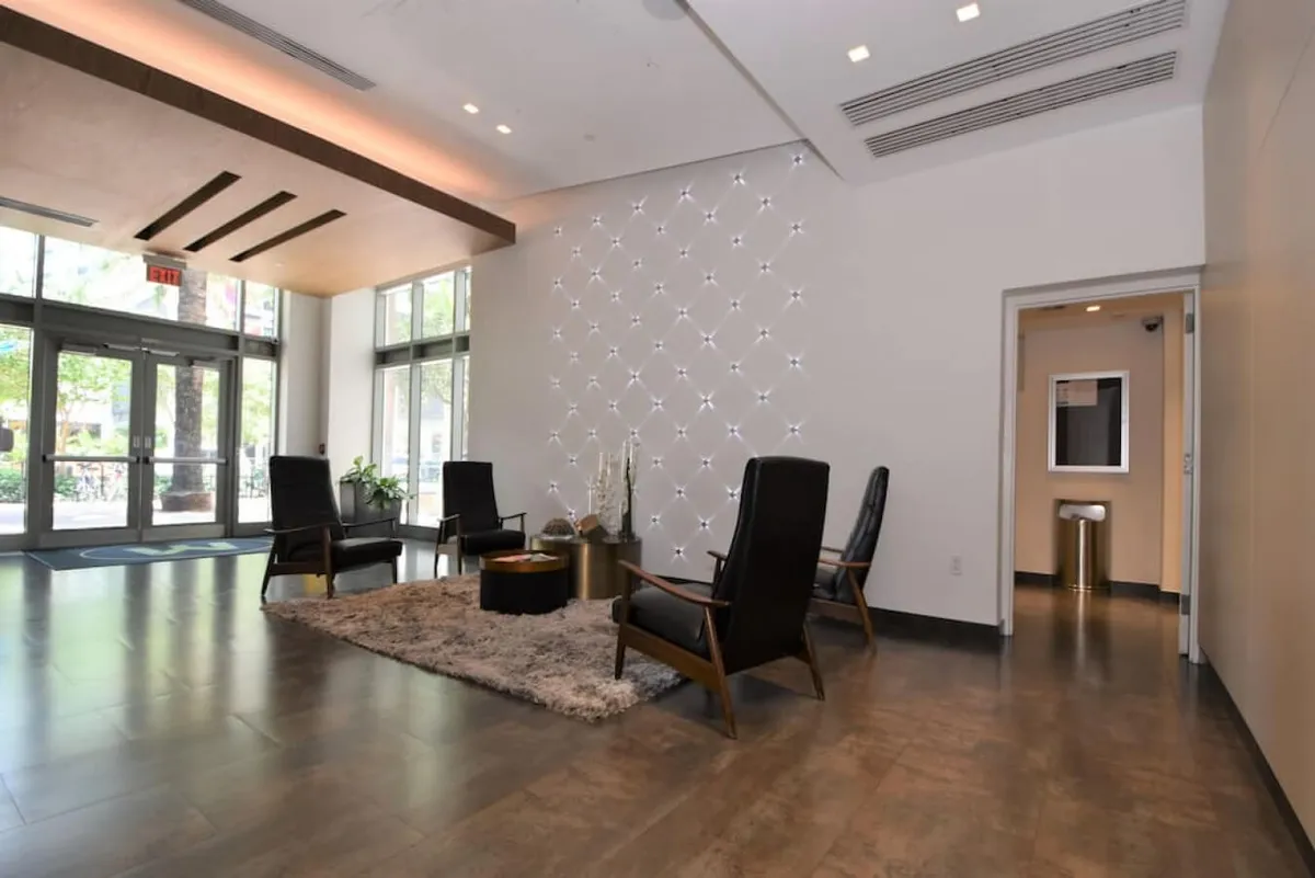 A welcoming lounge space featuring four black lounge chairs arranged around a low table, a decorative wall with an illuminated star-like pattern, and large glass doors letting in natural light. The space feels calm and inviting, ideal for relaxing or meeting guests, and is positioned near the building’s entrance and front desk.