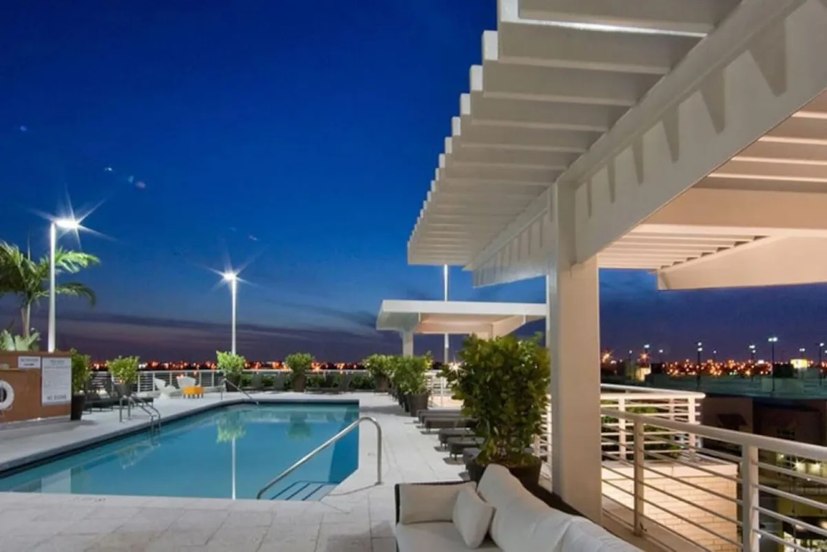 An illuminated swimming pool surrounded by comfortable outdoor seating with cushions, beneath a modern pergola structure. The backdrop features the city skyline with twinkling lights, creating a sophisticated and serene atmosphere perfect for evening gatherings. The scene conveys a luxurious, resort-style amenity that is central to the community experience.