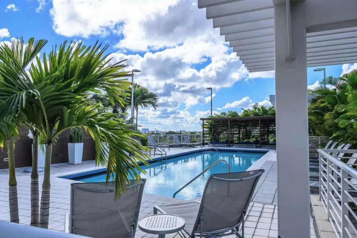 A rooftop swimming pool surrounded by lush tropical plants offers two lounge chairs and a shaded seating area under a pergola. The space provides sweeping views of the city, creating a tranquil oasis above the urban landscape.