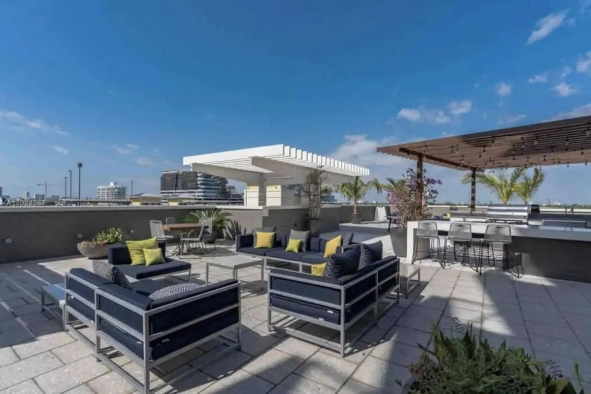 A modern rooftop outdoor seating area with navy blue cushions and yellow accent pillows, a bar-style counter with stools under a pergola, lush planters, and expansive city skyline views. The setup invites socializing, hosting gatherings, or relaxing in a stylish urban setting.